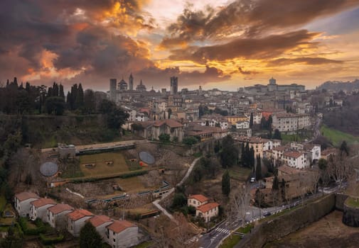 The old town of Bergamo, Italy, features medieval towers, domes, terracotta roofs, and lush greenery under a vibrant orange and purple sunset sky.