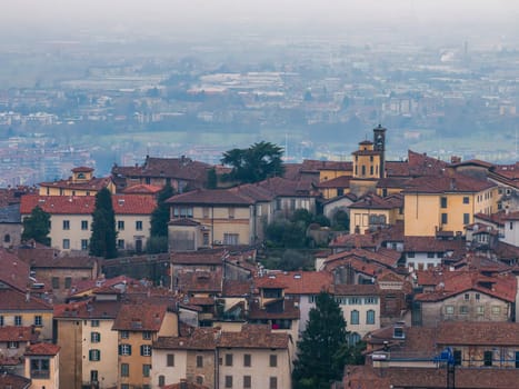 View of Bergamo's old town in Italy, featuring red tiled rooftops, a small bell tower, and a hazy backdrop of the modern city and countryside.