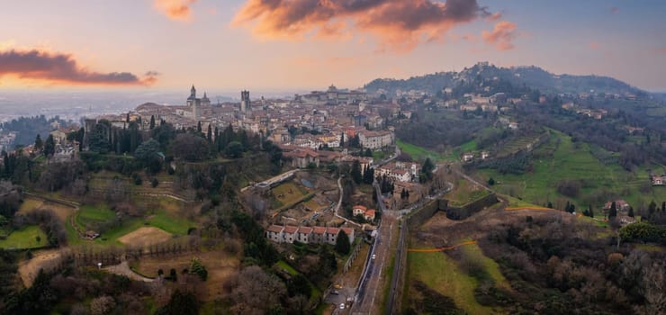 The old town of Bergamo, Italy, features medieval towers, domes, and Venetian walls, framed by green hills and bathed in warm sunset hues.