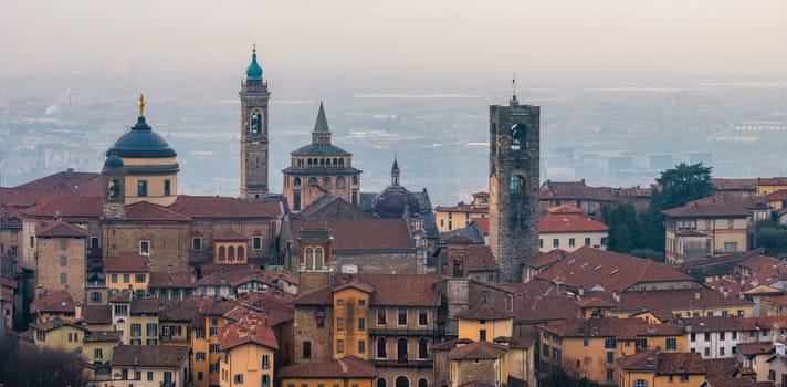View of Bergamo's old town featuring the Basilica of Santa Maria Maggiore, Campanone, and Cappella Colleoni amid terracotta roofs and a hazy sunset.