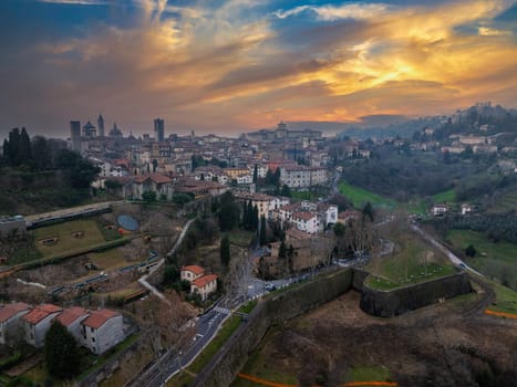 The old town of Bergamo, Italy, features medieval towers, domes, and stone walls, set against a vibrant sunset with rolling hills in the background.