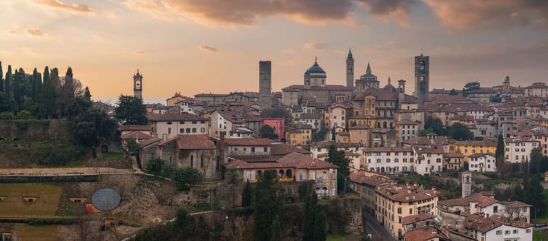 View of Bergamo's old town in Italy during sunset, featuring the Campanone, Basilica of Santa Maria Maggiore, medieval architecture, and lush greenery.