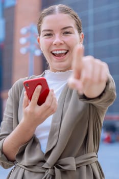 Smiling young woman with red smartphone pointing at camera, wearing casual outfit outdoors. Confident and cheerful expression, urban background. Perfect for lifestyle, social media, and advertising