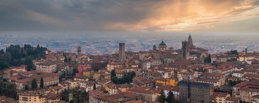 View of Bergamo's old town featuring the Basilica of Santa Maria Maggiore, the Campanone, red tiled rooftops, and a dramatic sunset sky.