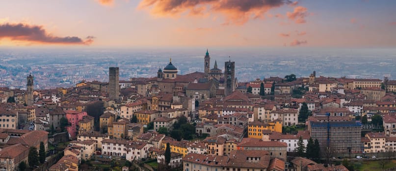 View of Bergamo's old town, Italy, at sunset, featuring the Basilica of Santa Maria Maggiore, medieval bell towers, and terracotta rooftops.