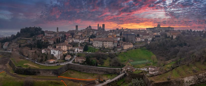 The old town of Bergamo, Italy, features medieval towers, domes, and Venetian walls under a vibrant sunset with pink, orange, and purple hues.