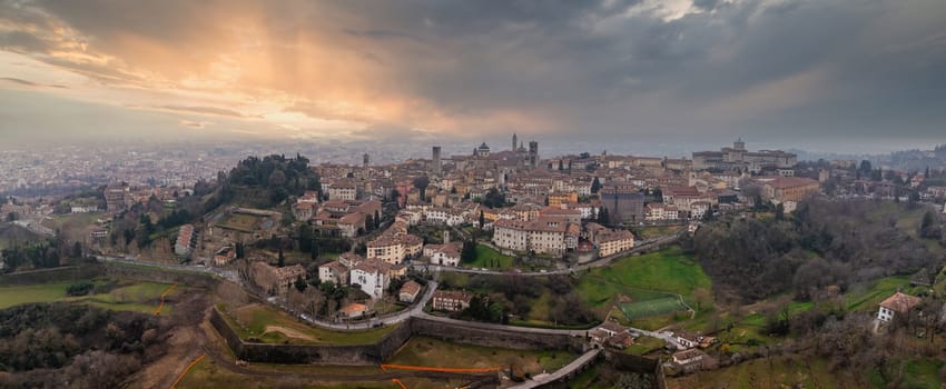 View of Bergamo, Italy, at sunset, featuring the historic old town on a hill, Venetian walls, Basilica domes, green fields, and a modern cityscape.