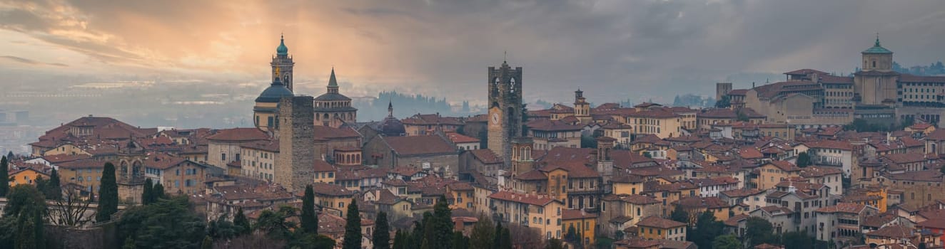 View of Bergamo's old town in Italy, featuring the Basilica of Santa Maria Maggiore, Torre del Gombito, red tiled roofs, and lush greenery at sunset.