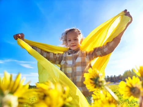 A child happily waves a bright yellow scarf while surrounded by blooming flowers in a sunny afternoon field.