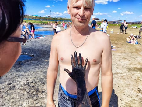 A middle-aged couple shares a lighthearted moment at a beach destination, with one partner playfully applying mud to the other. Their laughter fills the air as they enjoy their trip.