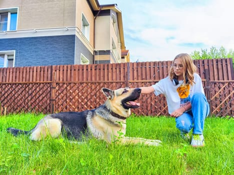 Girl sits on grass and gently pets her German shepherd dog in a residential backyard under clear blue skies.