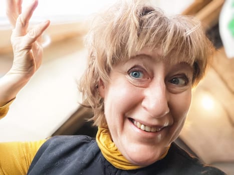 A middle-aged blonde woman smiles widely while taking a selfie indoors, sharing her vibrant travel experiences.