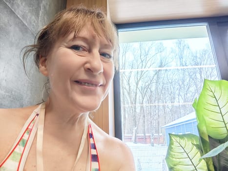 Mature woman takes a cheerful selfie near a window, surrounded by indoor plants, capturing a bright winter day outside.