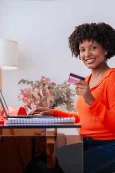 African american student making an online payment with a credit card while using a phone at home. Modern workspace with books, stationery, and stylish decor. E-commerce,digital transaction,and finance