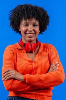 Smiling African American woman wearing red headphones,posing confidently with arms crossed against a blue background.Modern fashion, casual style, technology, self-assurance, and positivity.Copy space