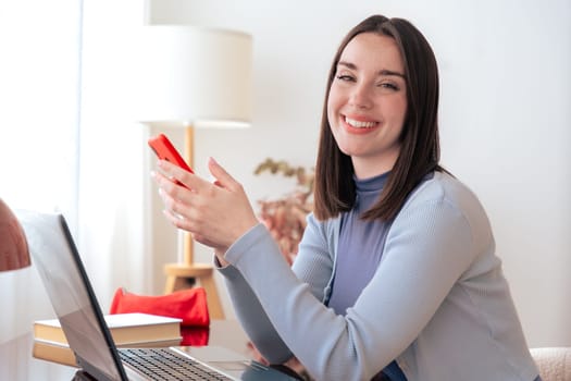 Happy Caucasian young woman looking at camera while working on laptop at home She holds a smartphone appearing confident and productive ideal for remote work freelancing or online communication themes