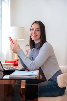 Happy Caucasian young woman looking at camera while working on laptop at home She holds a smartphone appearing confident and productive ideal for remote work freelancing or online communication themes