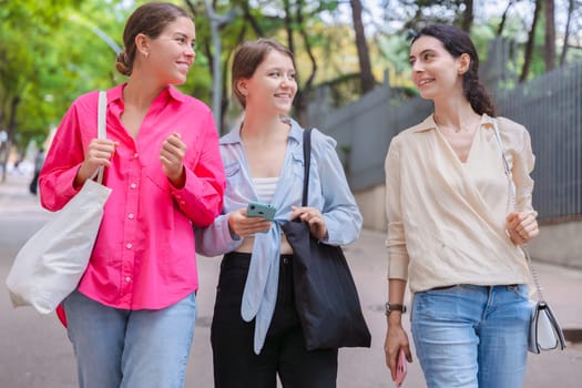 three gossipy teenagers chatting outdoors. A group of Caucasian friends chatting and communicating. Young adult students smiling and walking together at a social gathering.