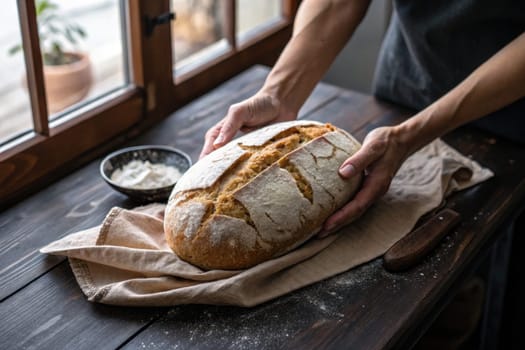 Warm, crusty bread is gently handled on a wooden table lined with a cloth. A small bowl of flour rests nearby, highlighting the process of baking.