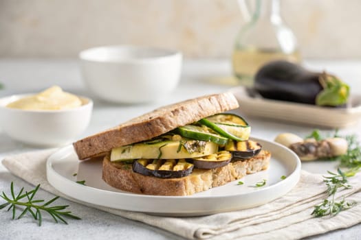 A grilled vegetable sandwich features layers of zucchini, eggplant, and green peppers on whole grain bread. It is garnished with herbs and served with a side of creamy dressing.