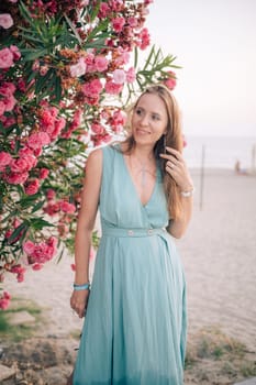 A graceful woman in a turquoise dress poses near blooming pink flowers, enjoying a peaceful moment in a natural outdoor setting.