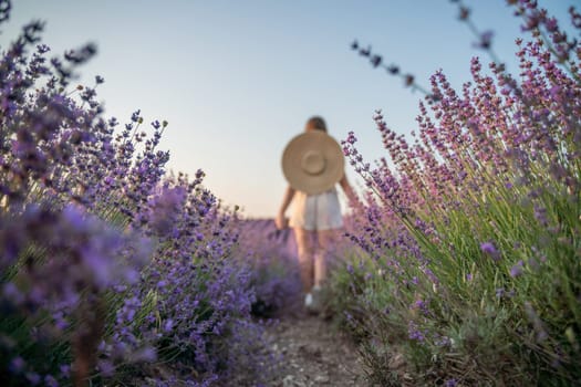A woman is walking through a field of purple flowers with a straw hat on.