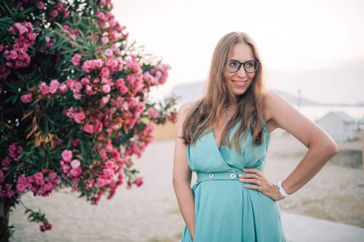 A graceful woman in a turquoise dress poses near blooming pink flowers, enjoying a peaceful moment in a natural outdoor setting.