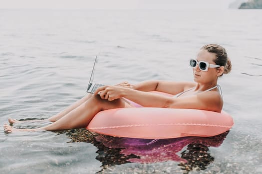 A woman is sitting in a pink inflatable raft on a lake, holding a laptop. Concept of relaxation and leisure, as the woman enjoys her time in the water while working on her laptop