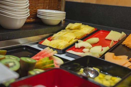 Indoor buffet with sliced pineapple, melon, watermelon, and kiwi in colorful trays, white bowls, and wicker basket on a dark countertop.