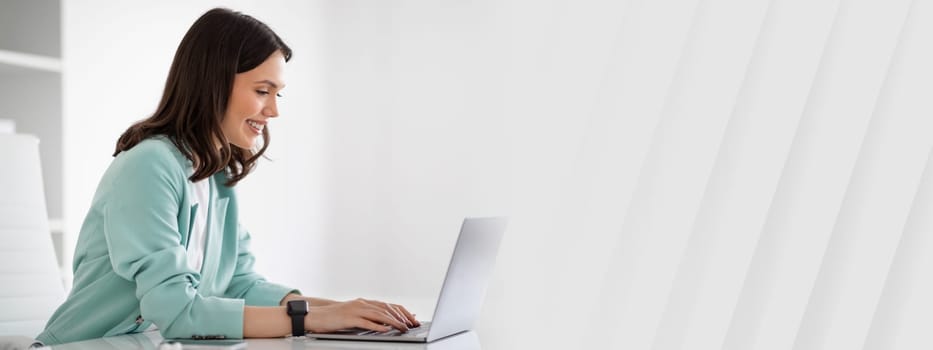 Smiling young european lady in suit at table typing on laptop, chatting with client in modern office interior, profile, ad, offer. Businesswoman and business, occupation, work, lifestyle with device