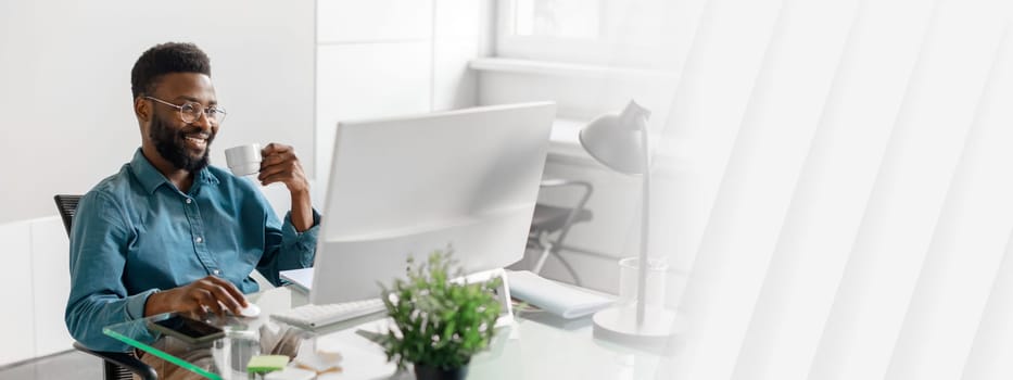 A young African American entrepreneur sits at a sleek desk, engaging with his work on a computer. He holds a cup, reflecting a positive lifestyle and managerial success during office hours.
