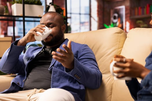Black couple sitting on the couch in their apartment and unwinding with warm drinks. Sharing an affectionate conversation and laughter that reflects happiness and companionship, love life.