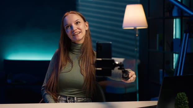 Girl in neon lit apartment using phone on selfie stick to capture welcoming intro for vlogging channel. Joyful entertainer in living room connecting with loyal audience during live stream, camera A
