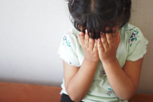 A young child sits quietly, covering her face with both hands indoors.