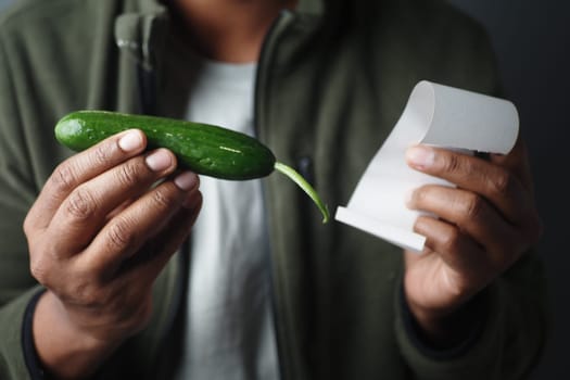 Man holds cucumber and receipt in indoor setting while examining them.