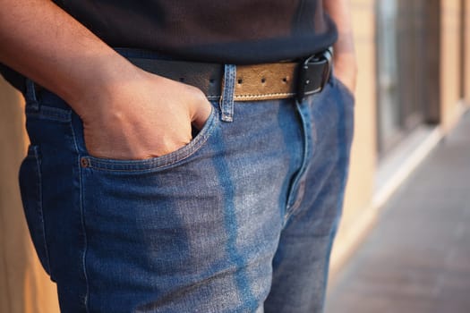 A person stands casually with hands in pockets, showcasing denim jeans and belt.