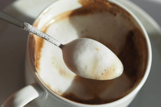 A spoon hovers over a cup filled with creamy coffee froth.