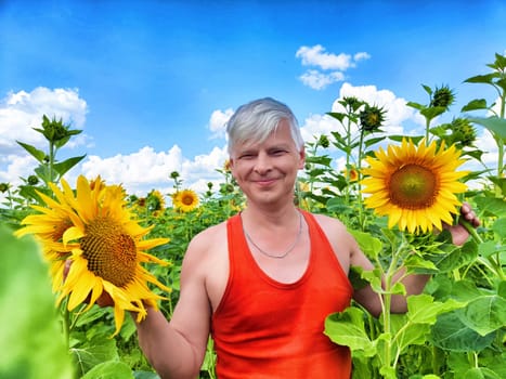 A man wearing a red tank top holds two large sunflowers in a lush field filled with blooming plants under a bright blue sky with fluffy clouds.