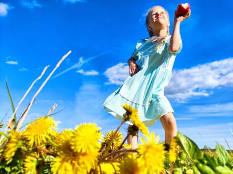 A girl in a blue dress is happily holding an apple amidst vibrant yellow dandelions and a clear sky, enjoying a sunny day outdoors.