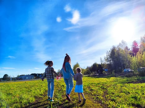 An adult and two children stroll together through a vibrant green field, enjoying a clear and sunny afternoon.
