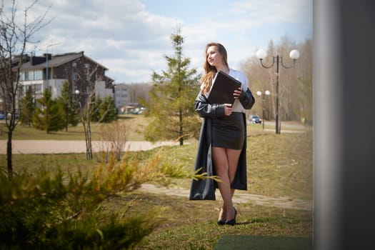A young woman with long hair stands confidently in a chic outfit, holding a folder near a building and trees.