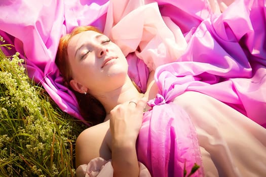 A woman enjoys a peaceful moment lying in a field, draped in bright pink fabric, absorbing the warm sunlight and nature around her.