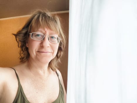 A cheerful woman with glasses stands by a window, enjoying the soft light filtering through sheer curtains.
