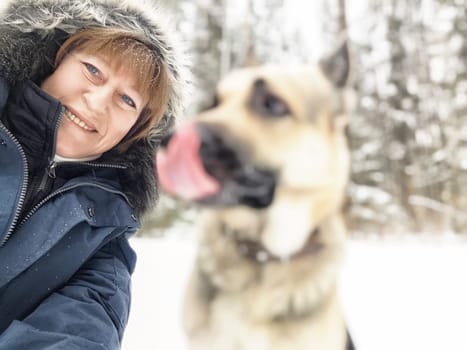A woman smiles while taking a selfie with her dog in a snowy landscape surrounded by trees.