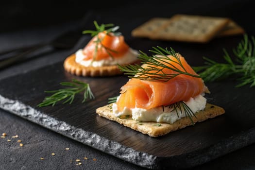 Two crispy crackers topped with smooth cream cheese and elegant slices of smoked salmon are garnished with fresh dill. This appetizer is arranged beautifully on a dark slate surface.