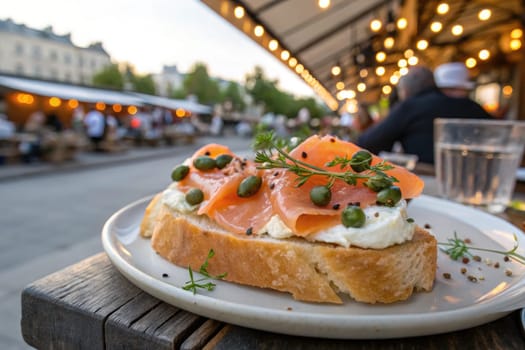 A plate of smoked salmon on toasted bread garnished with capers sits on a wooden table. Soft evening lights illuminate an outdoor dining area filled with people enjoying their meals.