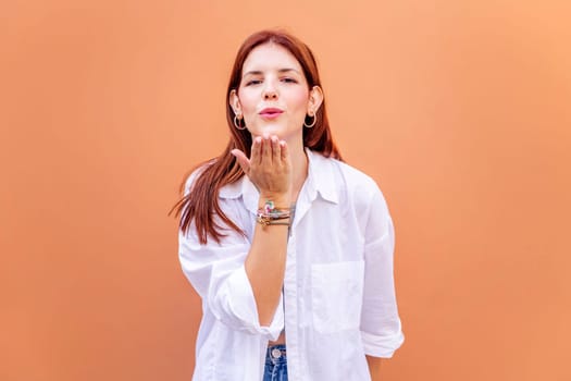 young woman giving a kiss with hand while smiling looking at camera in a positive and friendly mood, concept of love, copyspace for text