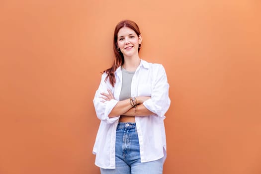 smiling young woman standing in front of orange wall looking at camera with her arms crossed, she is wearing a white shirt and blue jeans