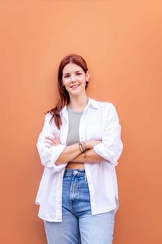 smiling young woman standing in front of orange wall looking at camera with her arms crossed, she is wearing a white shirt and blue jeans