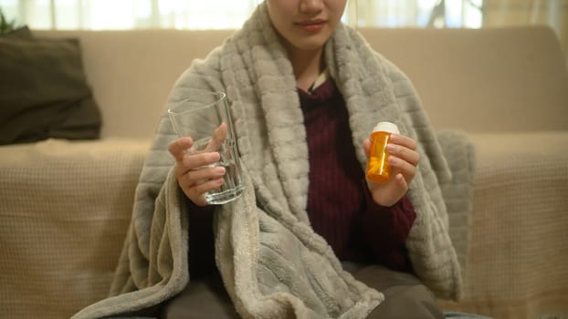 Sick young woman wrapped in a warm blanket and taking medicine for recovery.